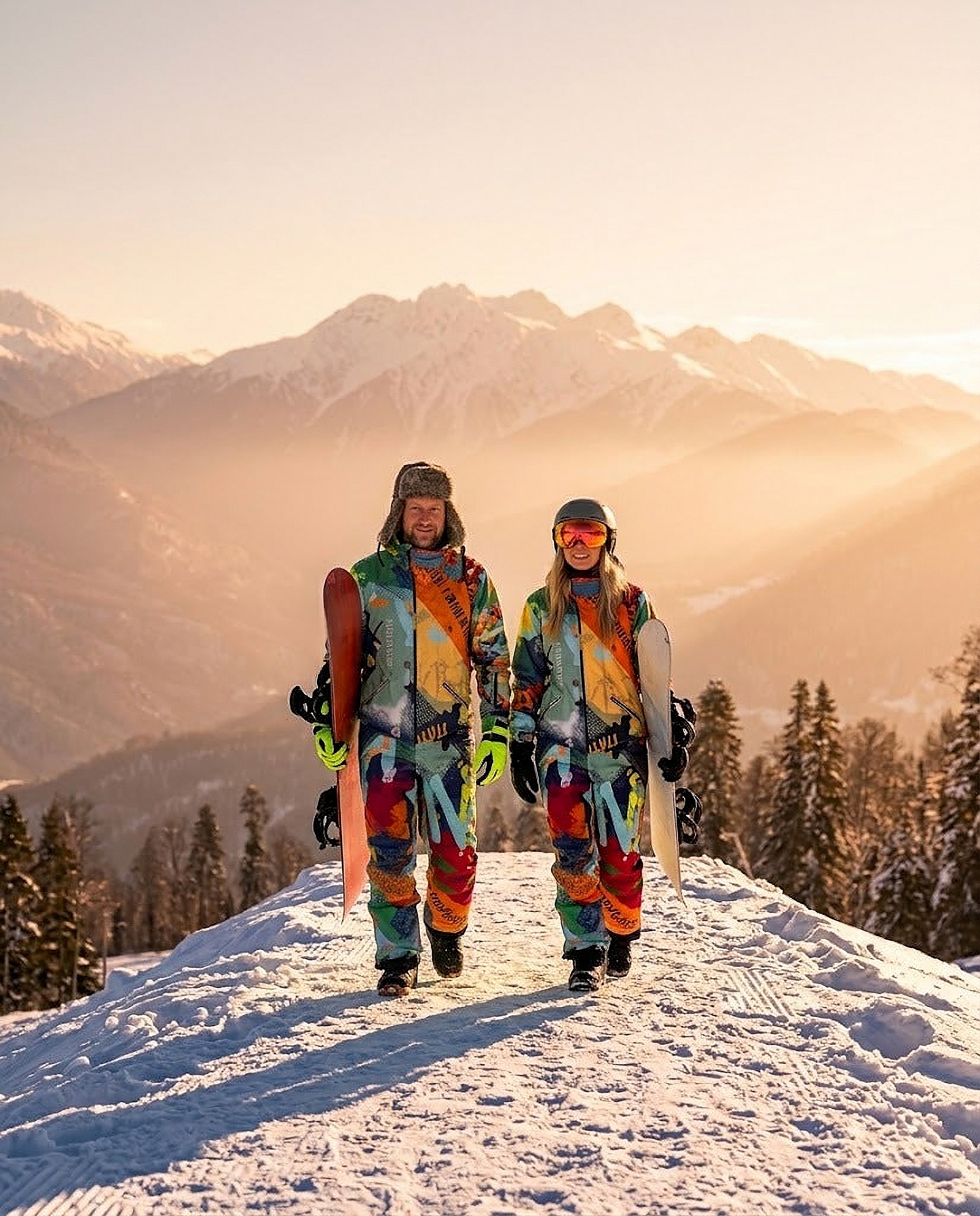 Couple in matching green multicolor print MoveAddict snowsuits carrying snowboards on a golden-hour alpine ridge